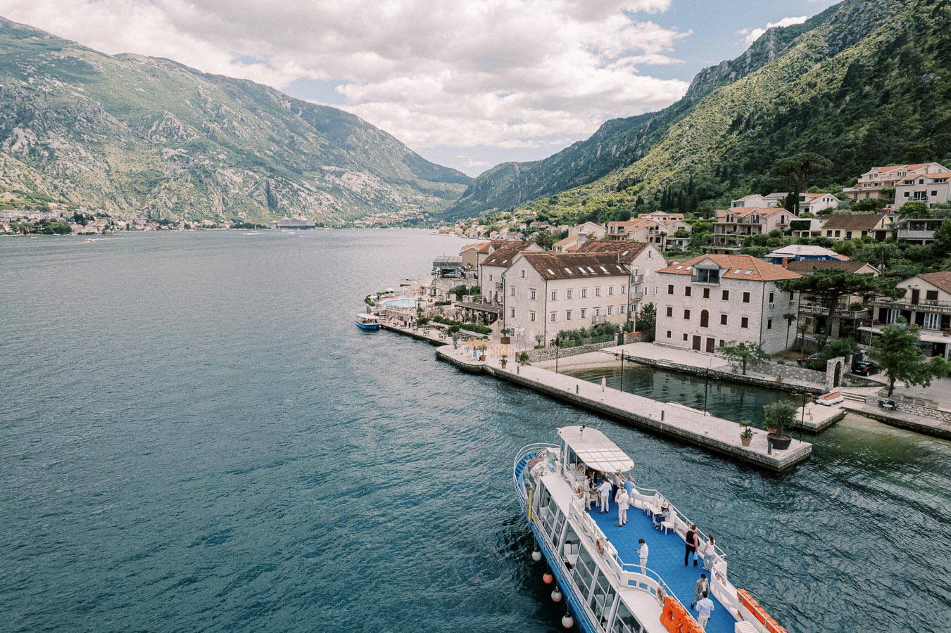 Scenic view of a coastal town with a boat in the Adriatic Sea, surrounded by lush green mountains under a partly cloudy sky, showcasing traditional European architecture.