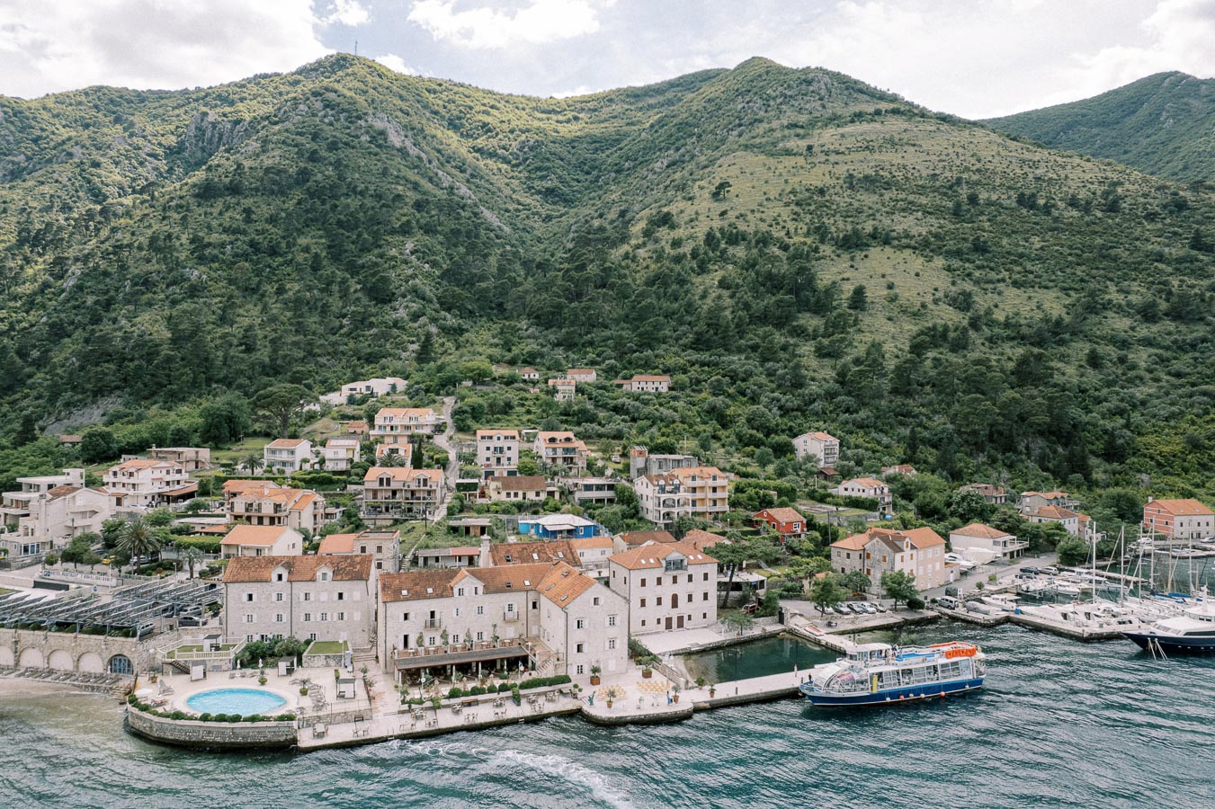 Aerial view of a coastal Mediterranean town with traditional stone houses and orange-tiled roofs, nestled against lush green hills, featuring a bustling marina with boats docked along the waterfront.