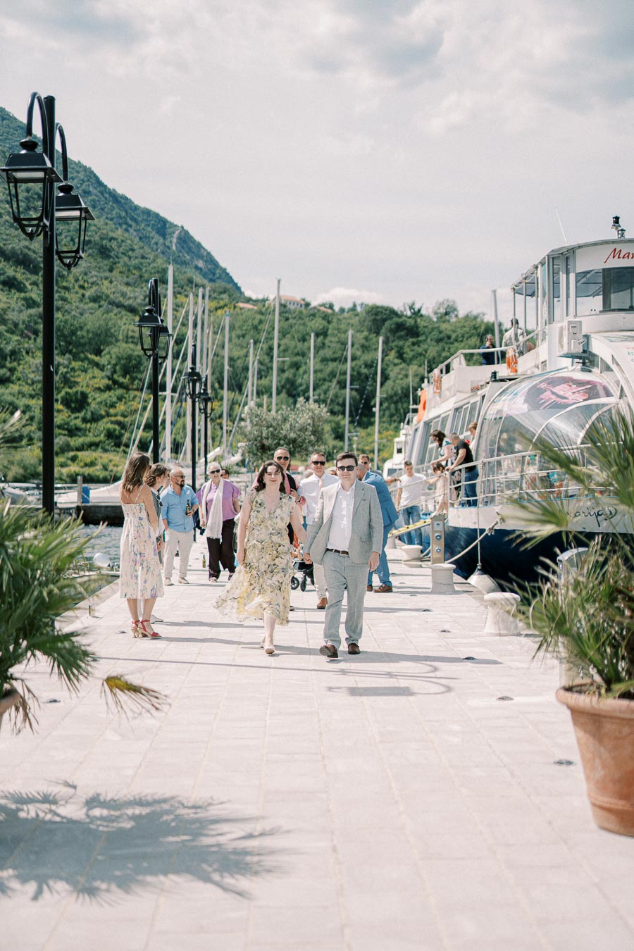 People walking along a sunny waterfront promenade by docked boats, with lush green hills in the background, under a partly cloudy sky.