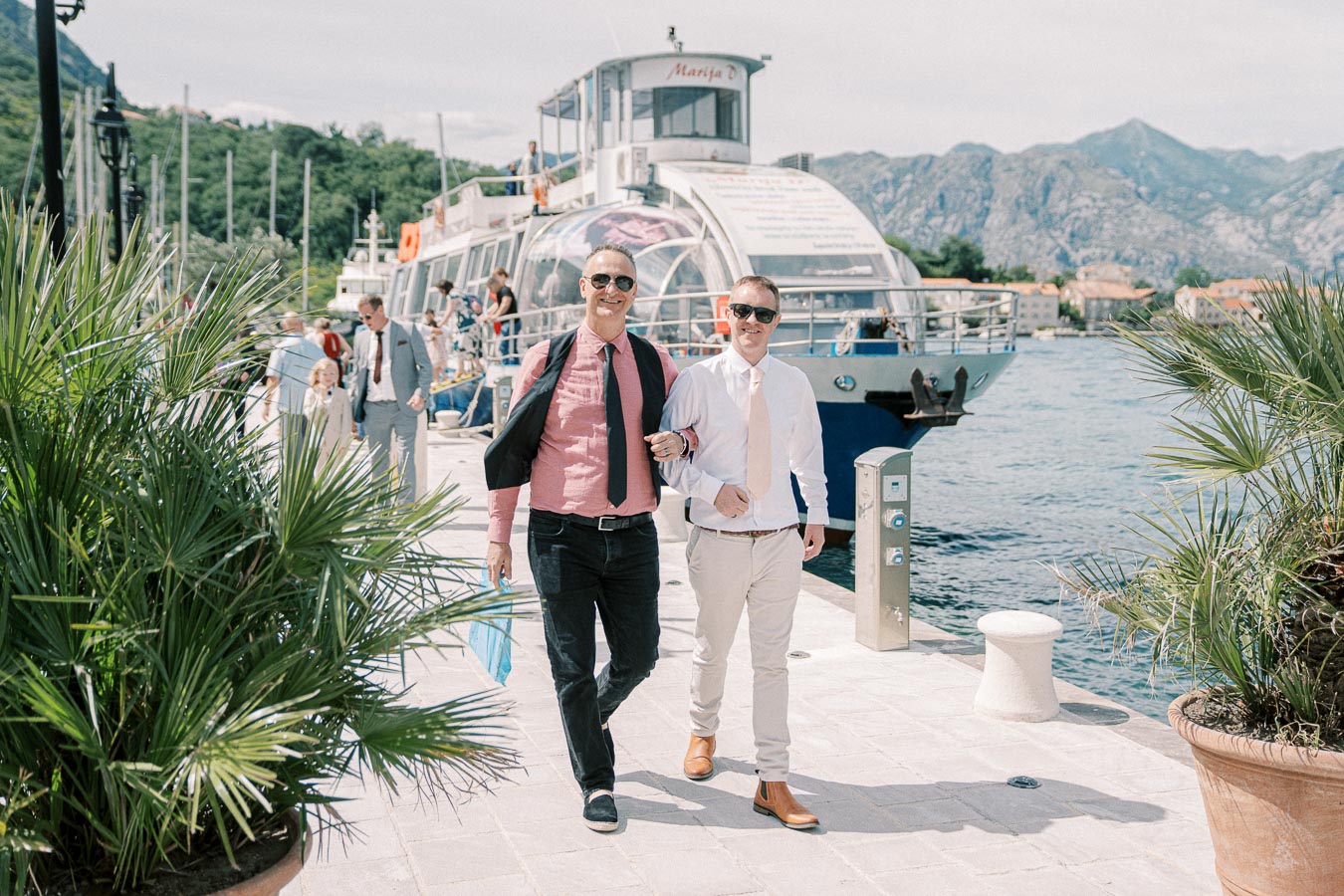 Two smiling men in sunglasses walking down a dock near a boat, surrounded by palm plants with a scenic mountainous background.