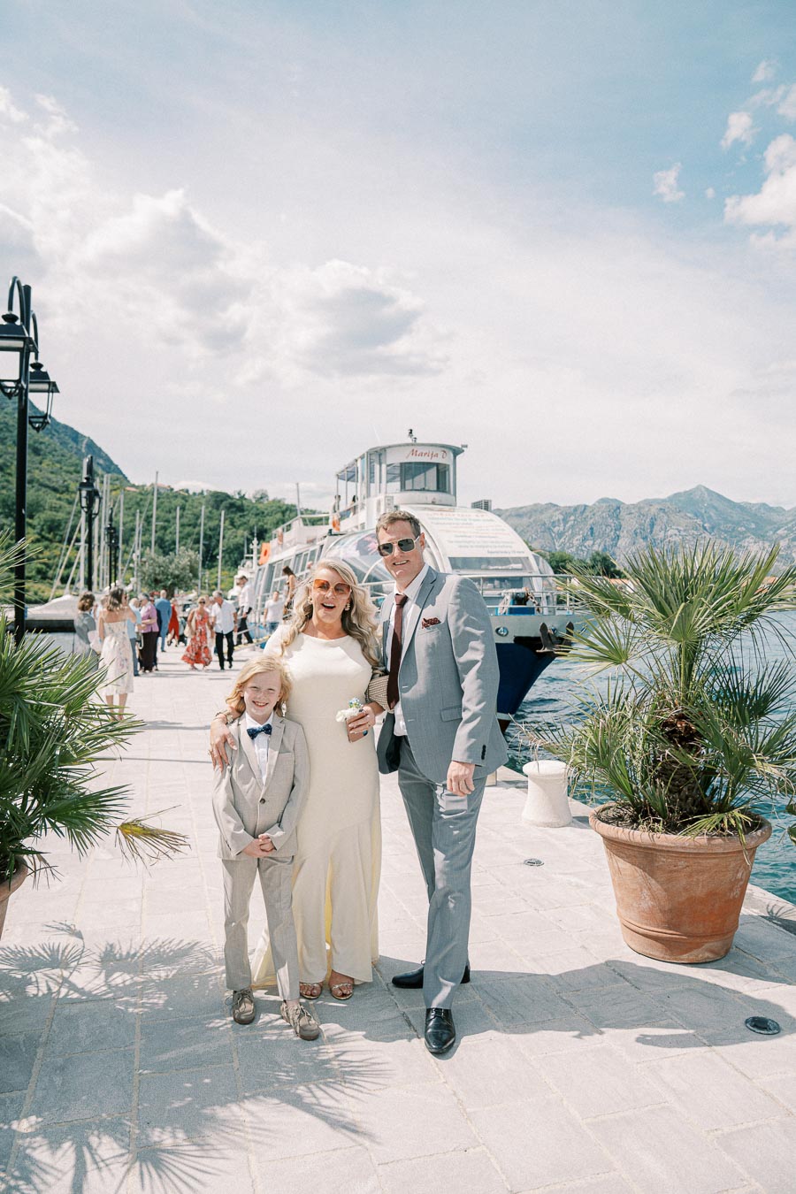 A family dressed in formal attire stands smiling on a scenic waterfront promenade with a boat docked behind them and mountains in the background, on a sunny day.