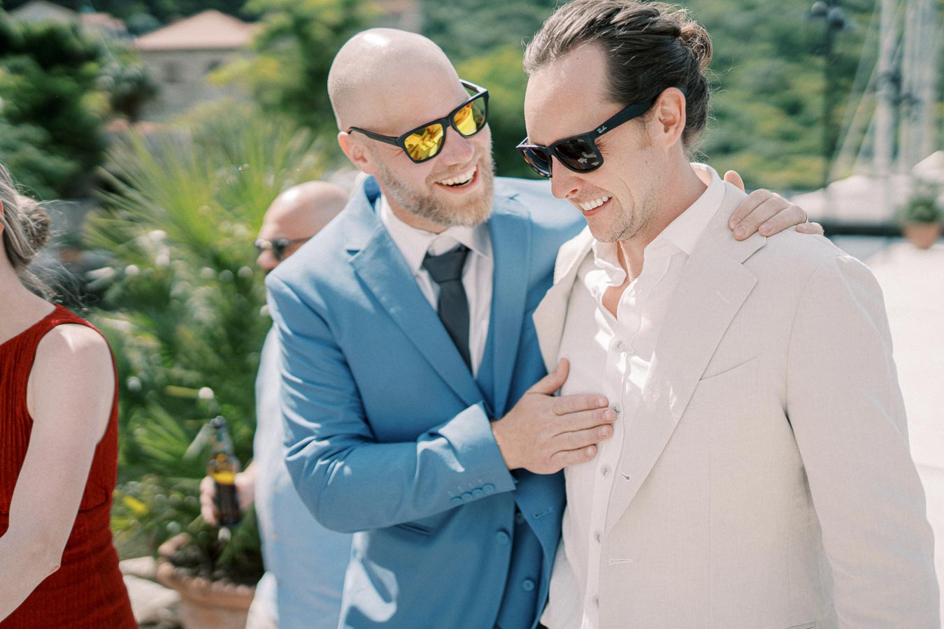 A group of people enjoying an outdoor event, with two men in suits and sunglasses sharing a cheerful moment, surrounded by greenery and relaxed ambiance.