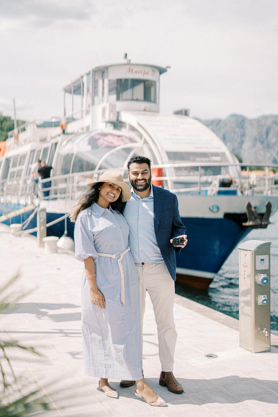 A smiling couple poses together on a sunny day by a docked boat, with mountains in the background, epitomizing a joyful vacation experience.