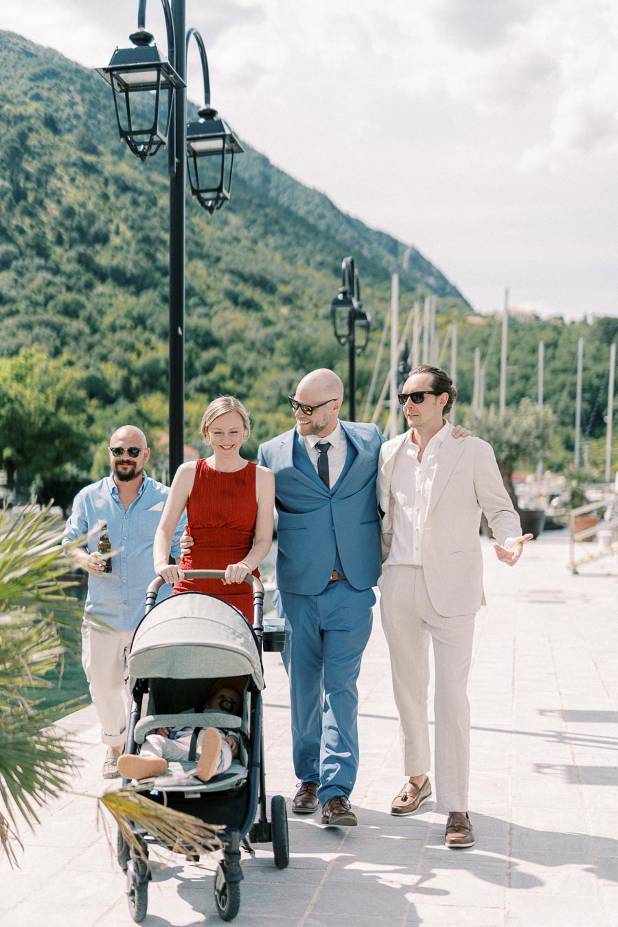 Group of well-dressed friends walking along a scenic promenade with a baby stroller and lush mountains in the background.