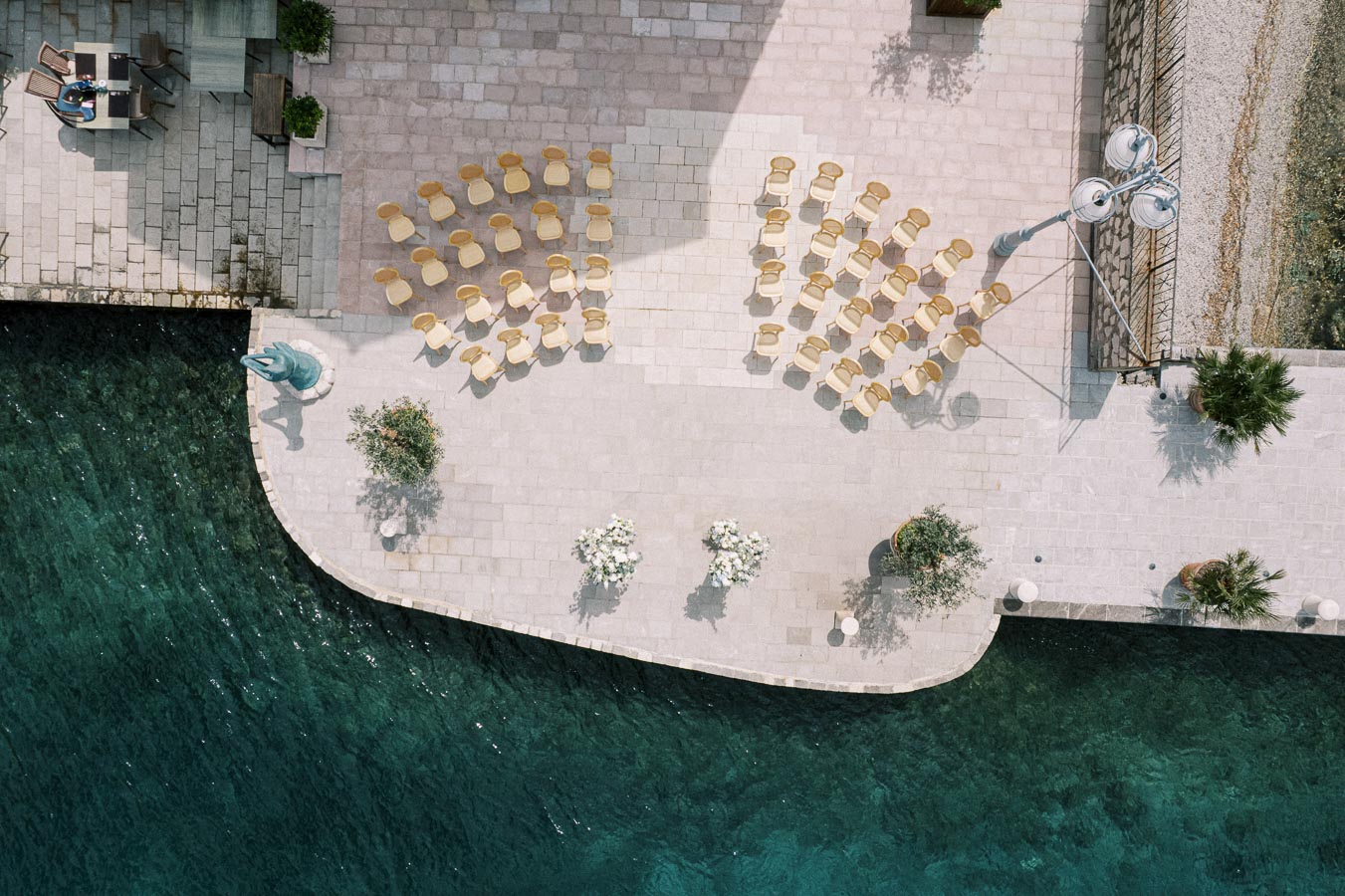 Aerial view of an outdoor event setup with neatly arranged wooden chairs on a stone patio near a waterfront, surrounded by potted plants and decorative elements, casting shadows on the ground and reflecting a serene, picturesque atmosphere.