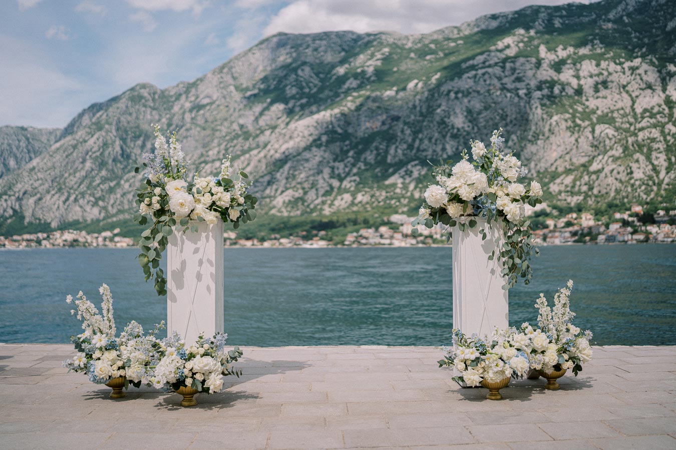 Scenic outdoor wedding ceremony setup with elegant white and blue floral arrangements by the waterfront, framed by majestic mountain views under a clear sky.