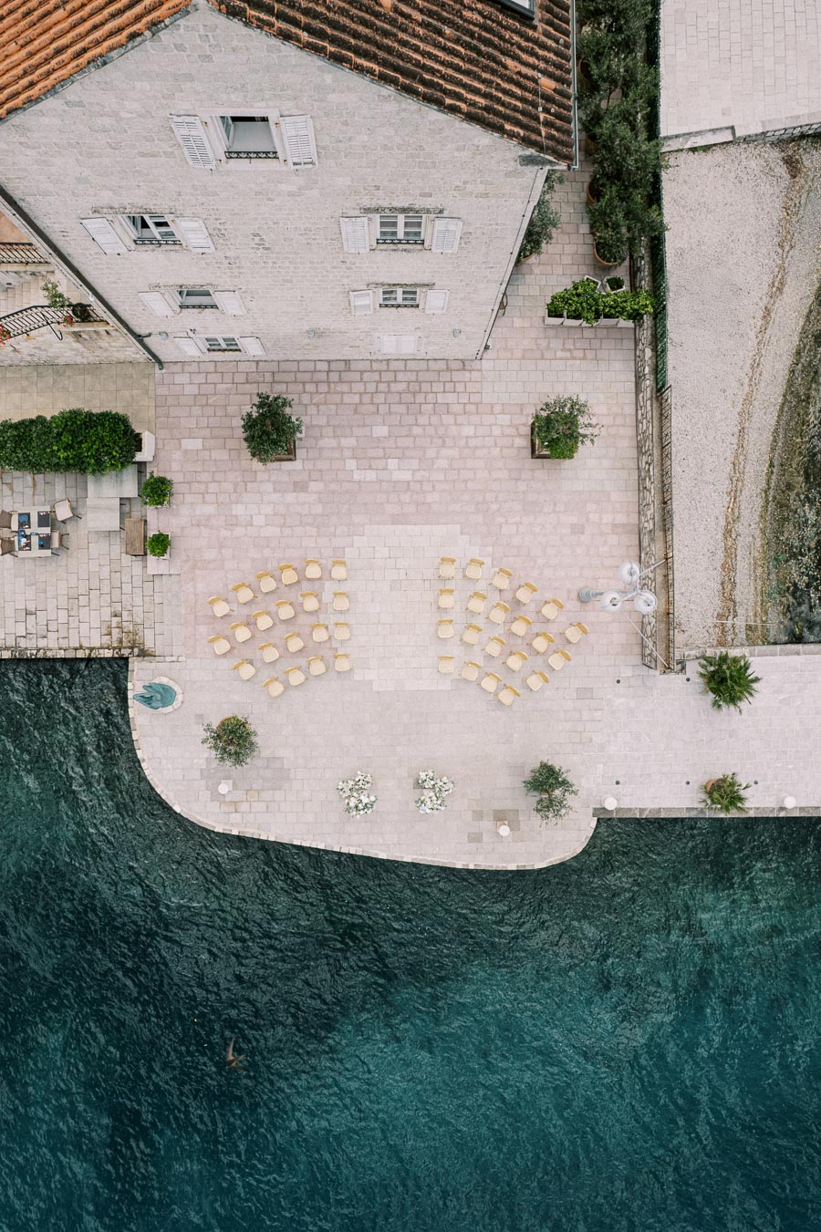Aerial view of a scenic Mediterranean waterfront terrace setup for an outdoor event, featuring arranged chairs, tables with floral decorations, and surrounding greenery, next to a historic stone building and vibrant blue water.