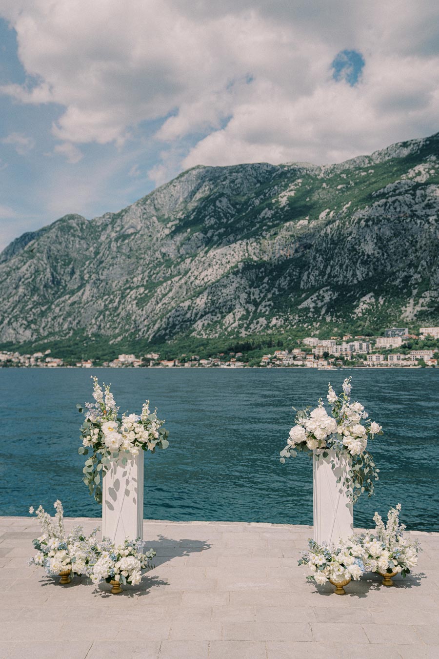 Outdoor wedding ceremony setup with floral decorations by a lake against a mountainous backdrop.