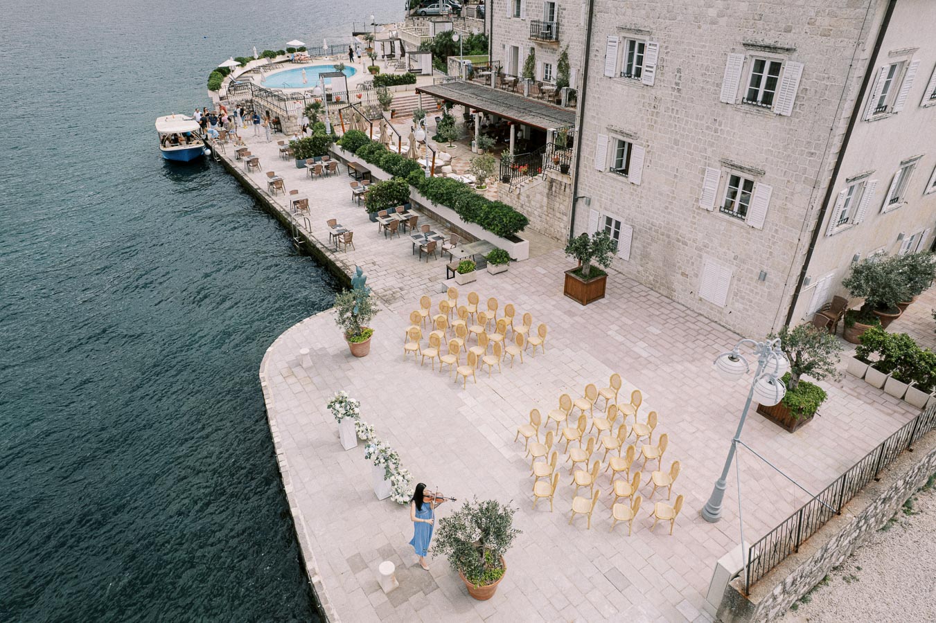 Aerial view of an outdoor waterfront wedding venue featuring arranged chairs, an elegant floral arch, and a musician playing a violin. The scenic backdrop includes a stone building and a calm body of water, emphasizing a serene and picturesque event setting.