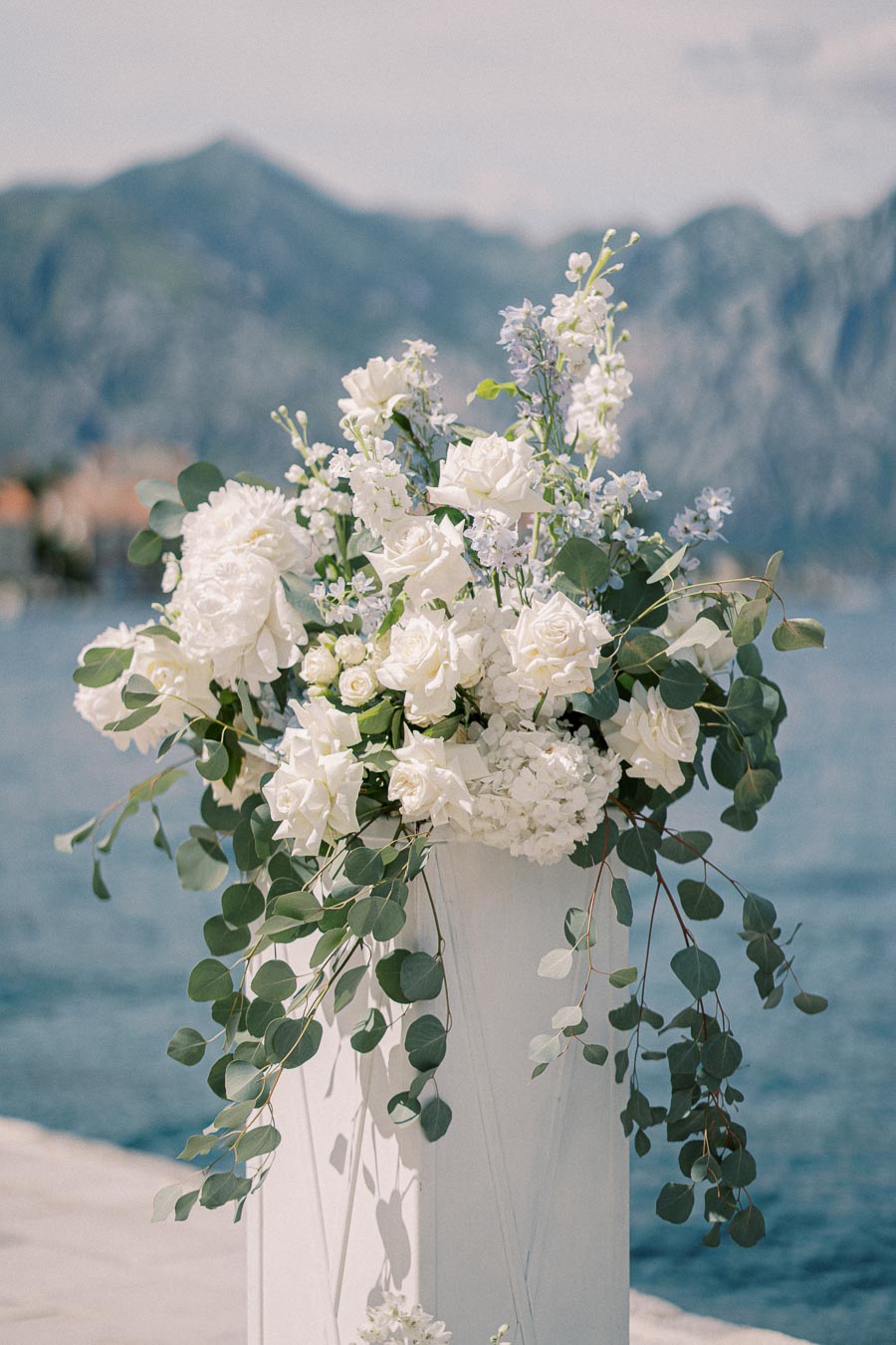 Elegant bouquet of white roses and greenery in a vase by the waterfront, with mountains in the background.