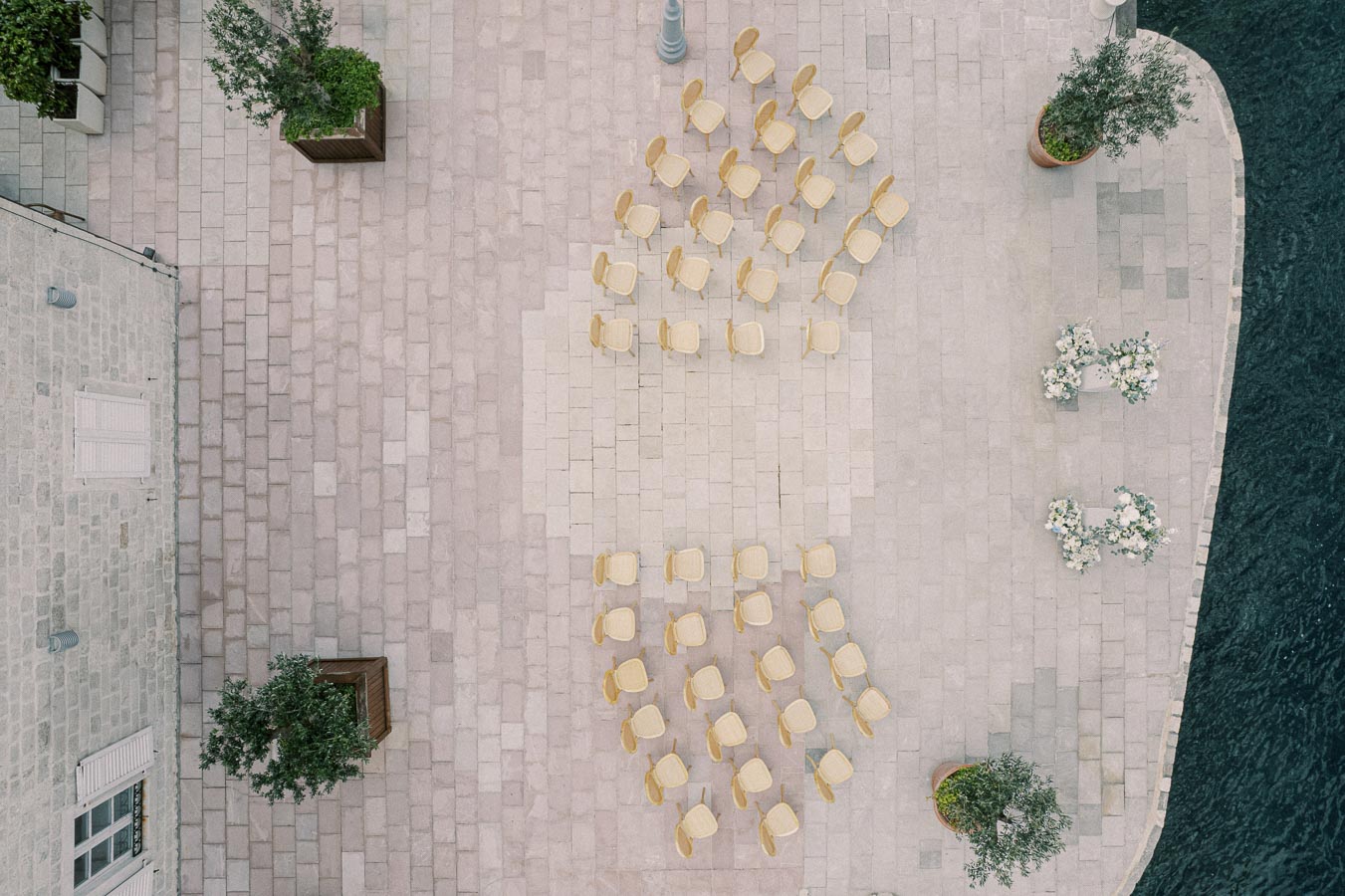 Aerial view of an outdoor venue setup for a wedding ceremony by the waterfront, featuring neatly arranged wooden chairs on a stone patio, adorned with white floral decorations and greenery.