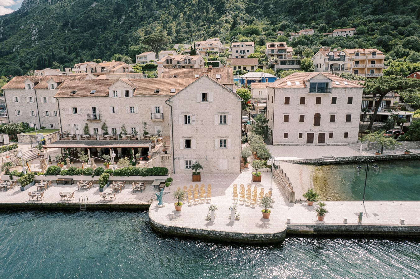 A picturesque coastal village featuring historic stone buildings with red-tiled roofs along a serene waterfront, surrounded by lush green hills. Empty outdoor seating and neatly arranged chairs suggest a tranquil setting for events or dining by the water.