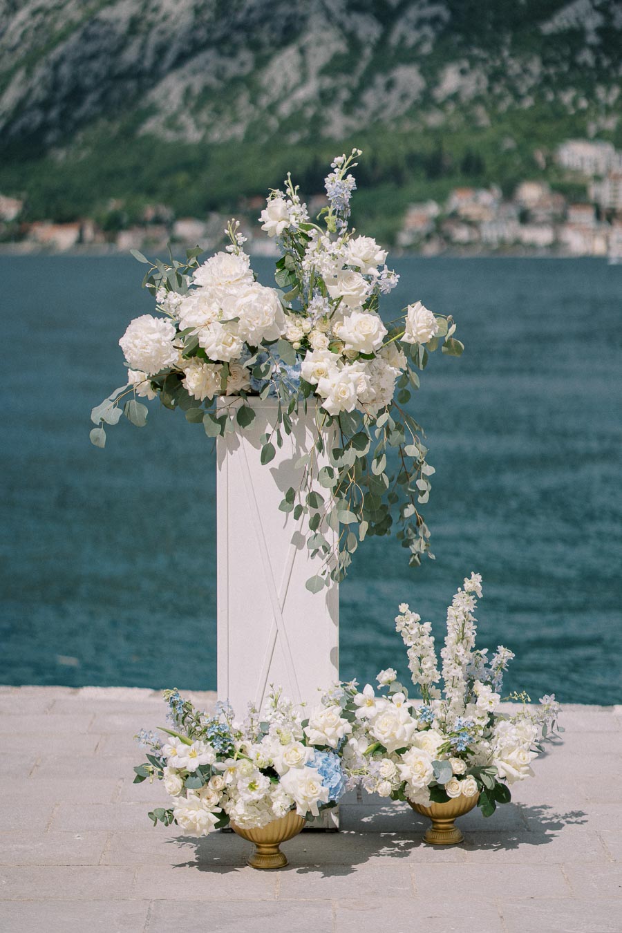 Elegant floral arrangements featuring white and blue flowers in ornate vases set against a scenic backdrop of a serene lake and lush mountains, perfect for outdoor weddings or events.