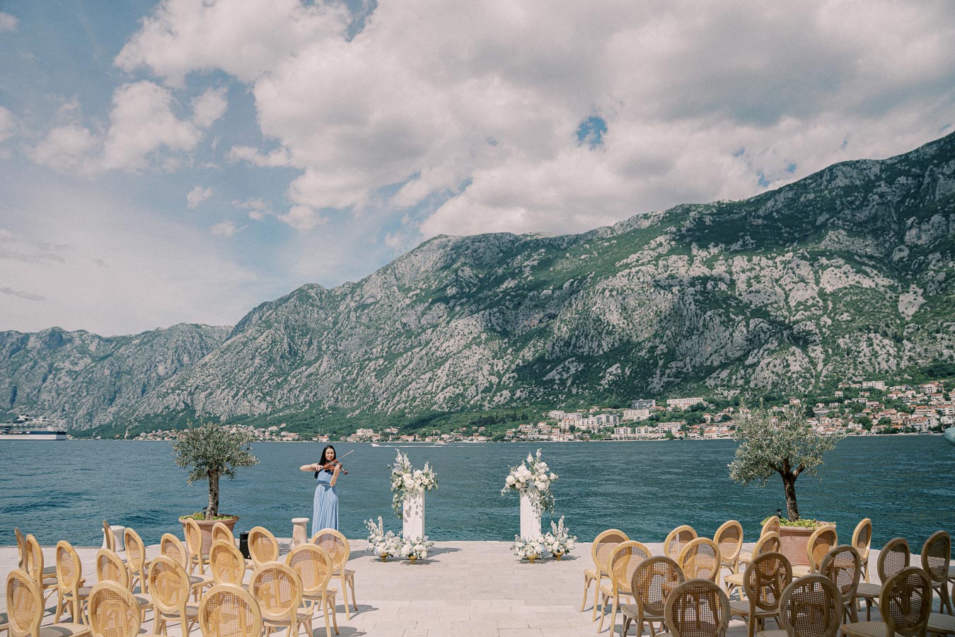 Wedding ceremony setup by a serene lake with majestic mountain backdrop, featuring elegant chairs, floral arrangements, and a violinist in a blue dress.