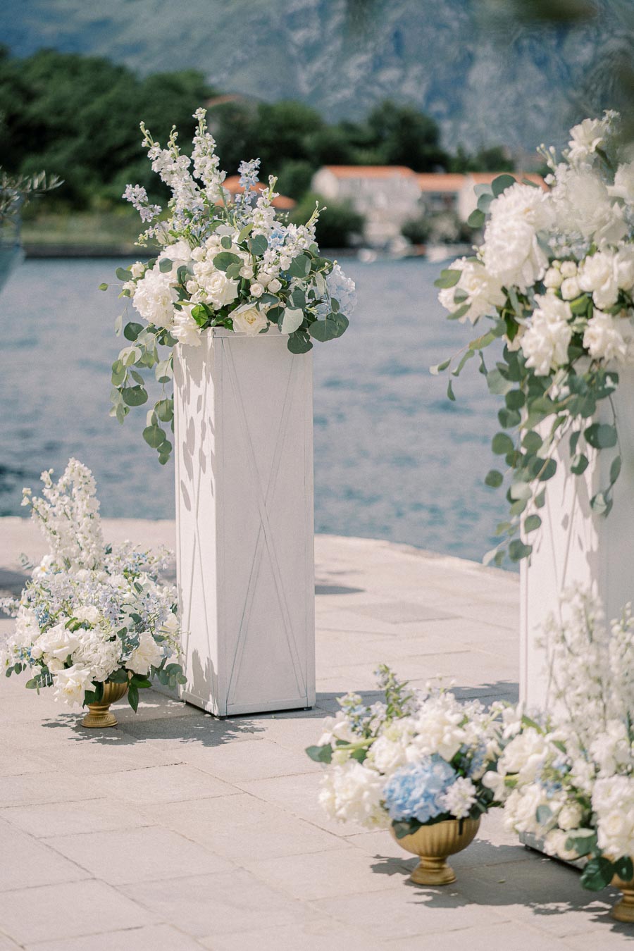 Elegant floral arrangements with white and blue flowers in tall white vases, set against a serene waterfront backdrop with distant mountains and lush greenery. Perfect setting for an outdoor wedding or event.