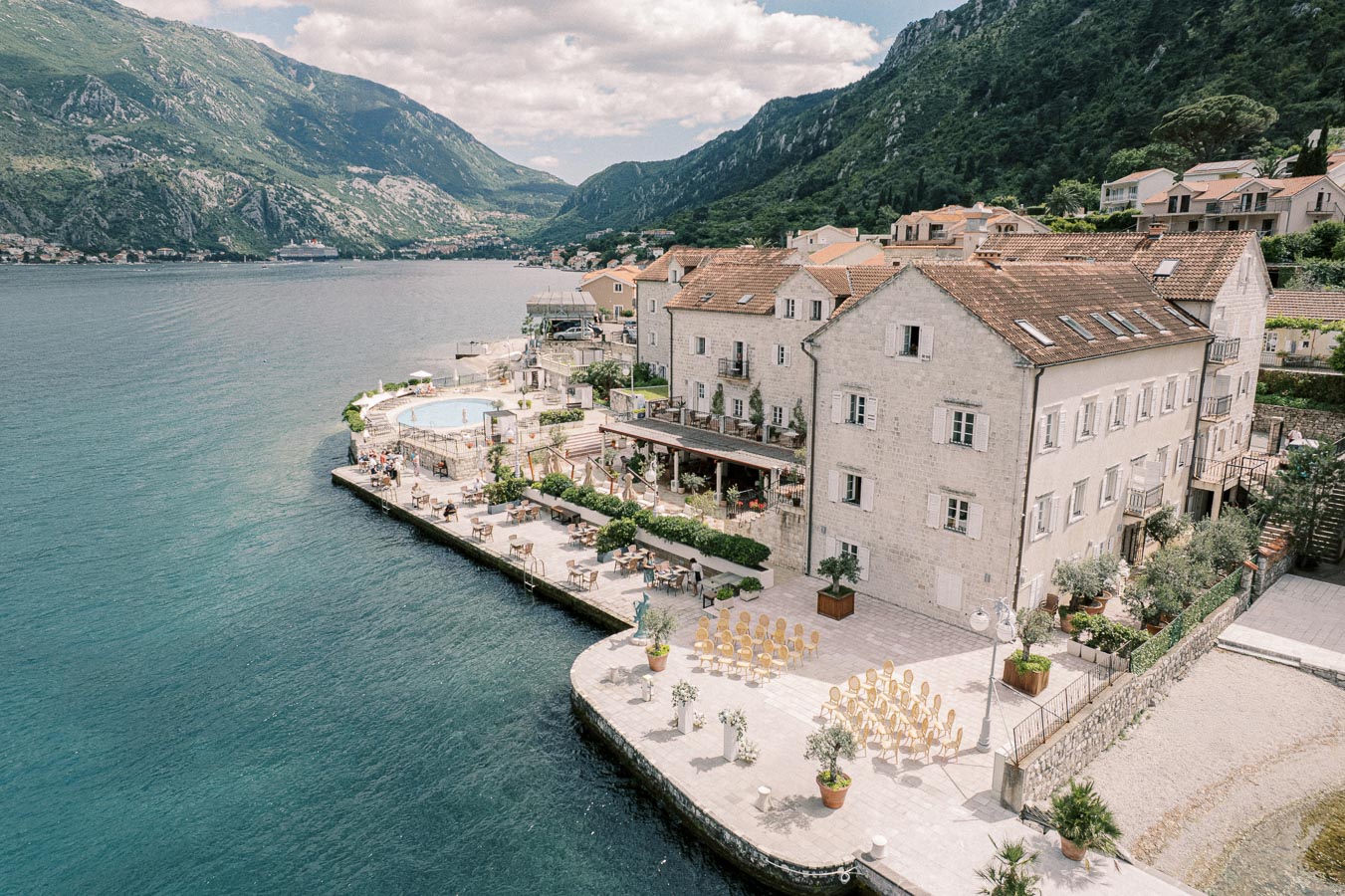 Aerial view of a picturesque seaside resort with elegant stone buildings, a pool area, and outdoor seating overlooking a calm bay surrounded by lush green mountains. Perfect destination for a peaceful getaway or special events.