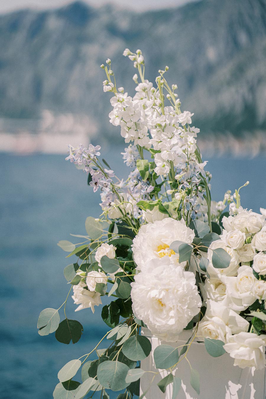 Beautiful floral arrangement with white and pastel flowers, including peonies and roses, set against a serene mountain and water backdrop, perfect for a wedding or outdoor event.