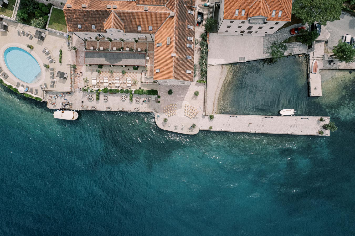 Aerial view of a waterfront resort with a swimming pool, outdoor seating, and boats docked at a pier, set against a backdrop of clear blue water.