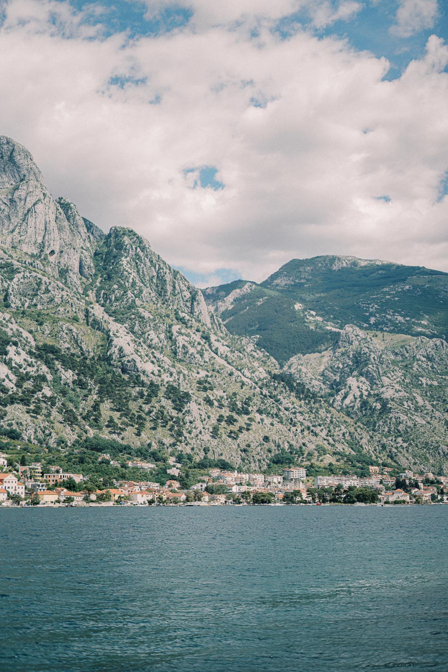 A scenic view of a coastal town beneath majestic, rocky mountains, with clear blue water in the foreground and a partly cloudy sky above.