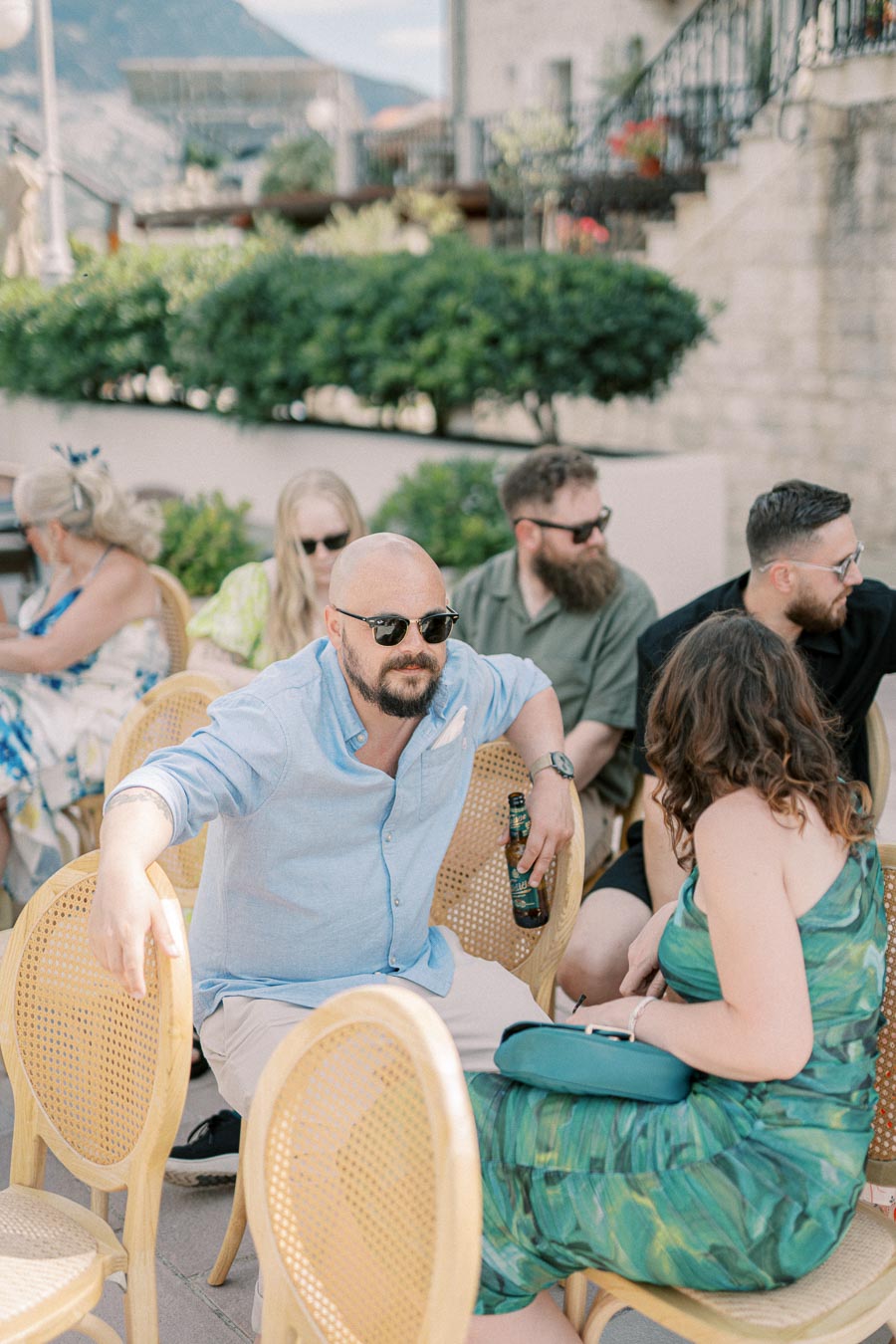 Group of people enjoying a casual outdoor gathering, with a man in a light blue shirt holding a beer and conversing with a woman in a green dress, set against a backdrop of greenery and stone buildings.