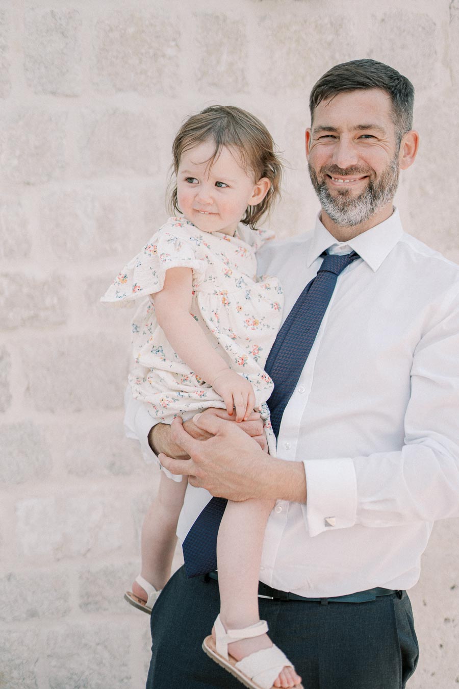 Father in a white shirt holding his daughter in a floral dress, standing against a stone wall background, both smiling warmly.