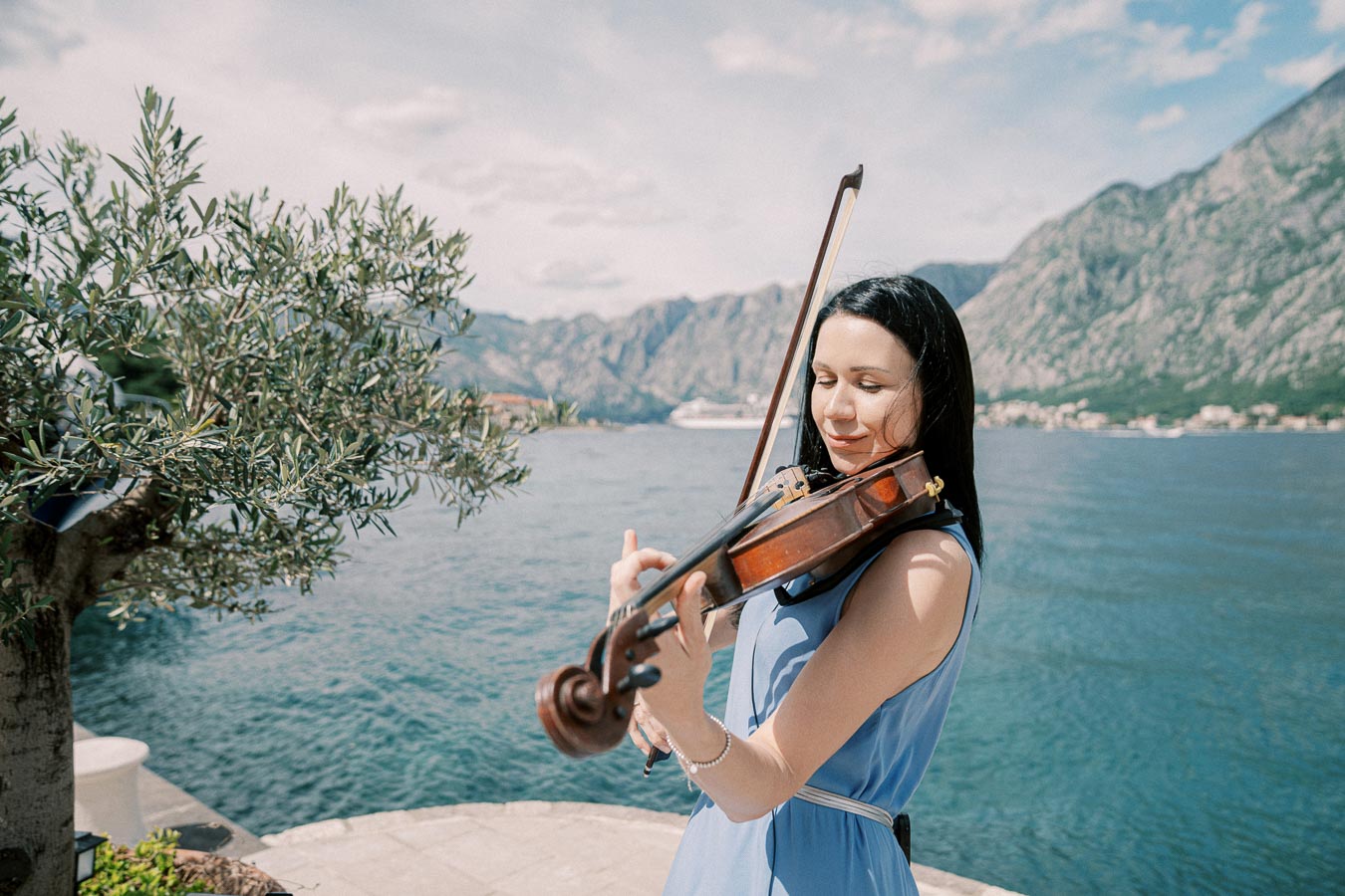 A woman in a blue dress plays the violin by a scenic waterfront, surrounded by mountainous landscapes and a clear sky.