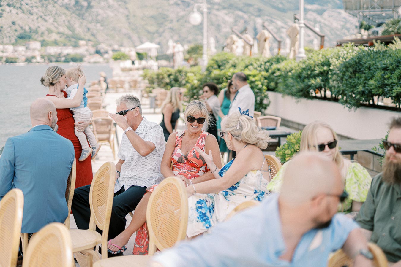 A group of people enjoying a sunny outdoor gathering by the water, surrounded by lush greenery and mountains in the background. They are seated on wicker chairs, conversing and wearing summer attire, while a woman in red holds a child.