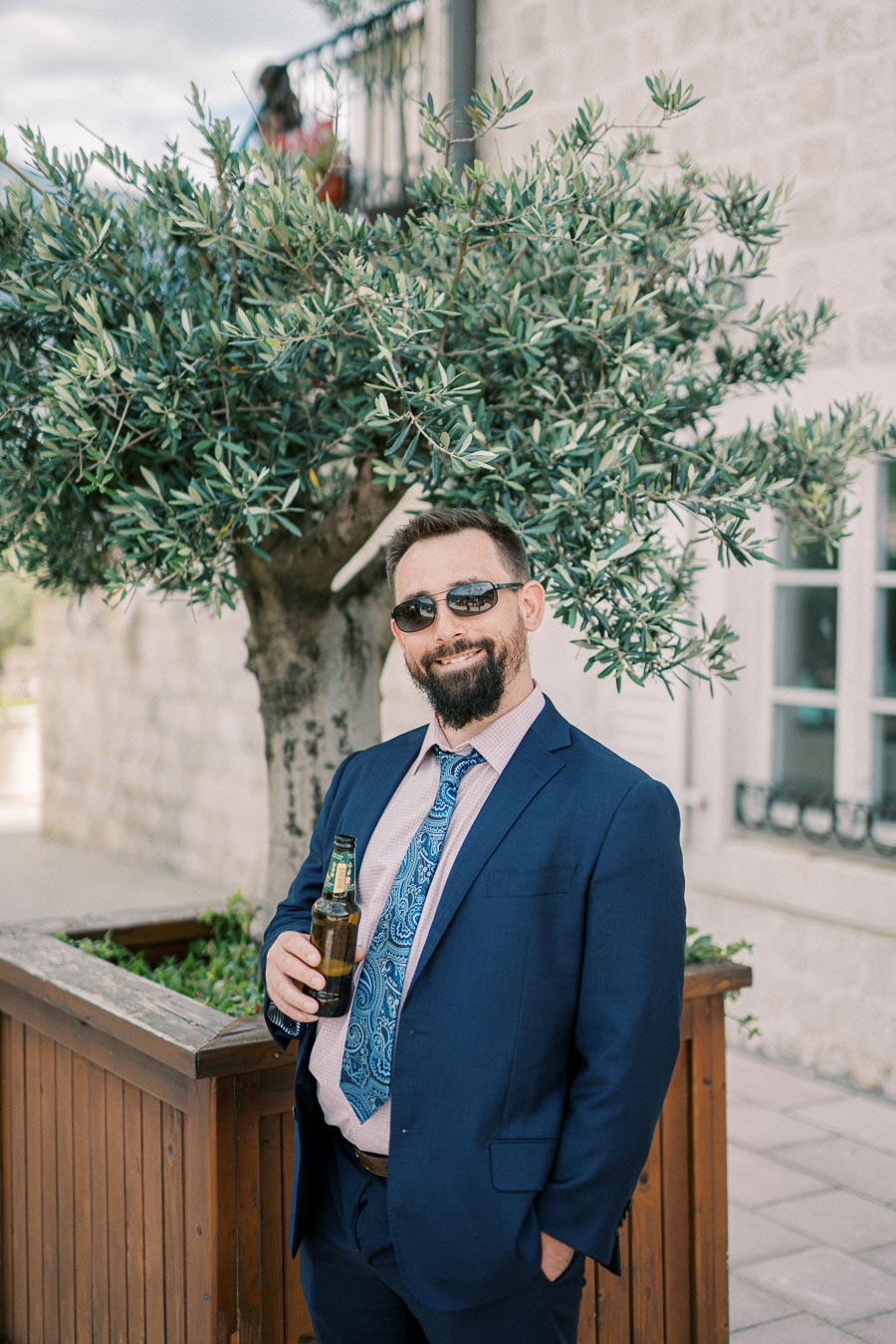 A man in a blue suit with sunglasses holding a beer bottle, standing next to a large potted tree outside a building with stone walls.