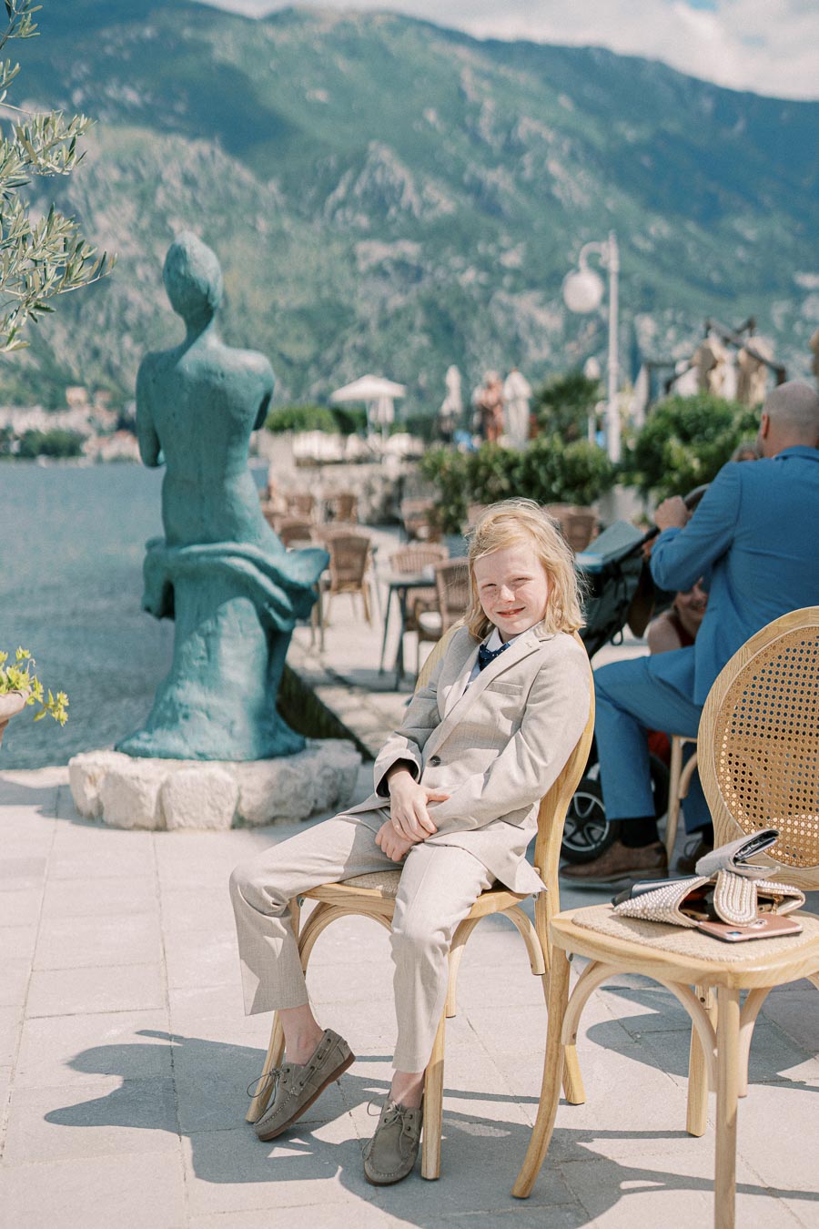 Young person sitting in a chair wearing a light-colored suit, with a scenic waterfront background including a bronze statue and mountainous landscape.