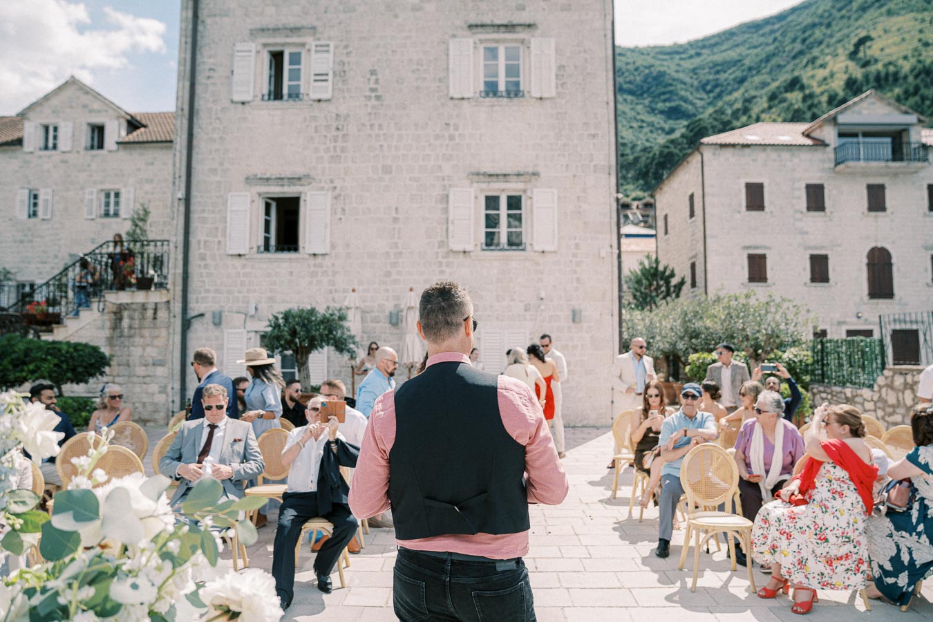 A wedding ceremony set outdoors with guests seated in front of a historic stone building. A man in a vest stands with his back to the camera, likely making an announcement. The scene is surrounded by lush greenery and mountainous scenery under a clear blue sky.