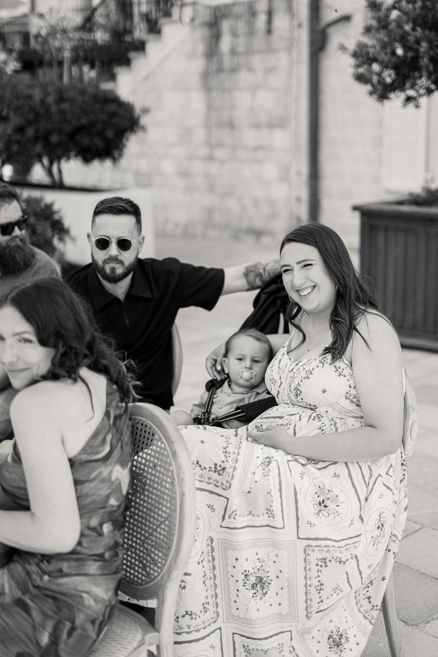 A black and white photo of a group of people sitting outdoors, featuring a woman smiling and holding a child on her lap while a man with sunglasses sits next to them.