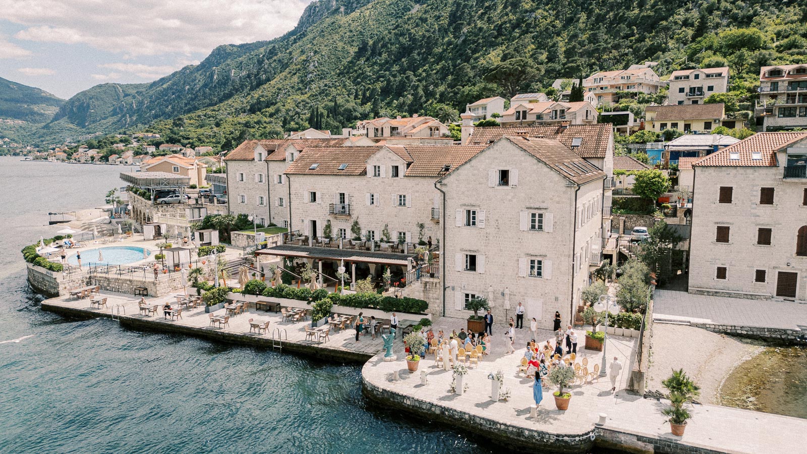 Aerial view of a picturesque coastal town with rustic stone buildings, a waterfront terrace with tables and seating, and a scenic backdrop of lush green hills and waterfront.