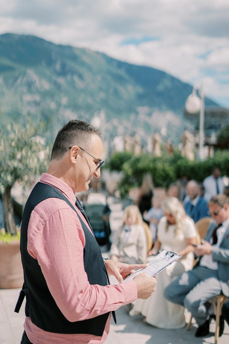 Man reading from a clipboard at outdoor wedding ceremony with scenic mountainous background, guests seated in the sunlight.
