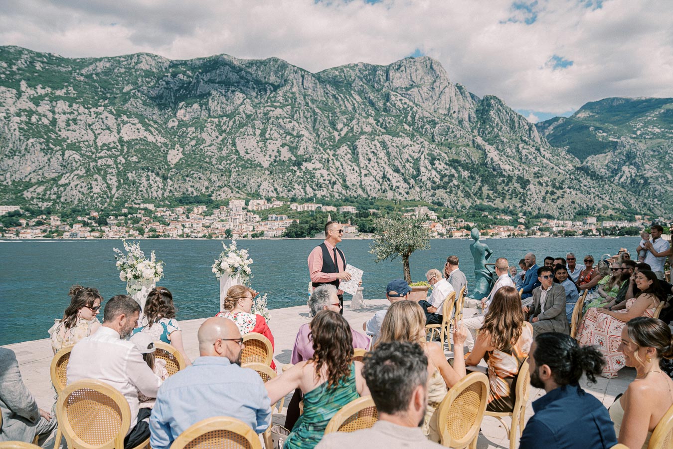 Outdoor wedding ceremony by a picturesque lake with mountains in the background, featuring guests seated on wicker chairs, surrounded by floral arrangements and a statue, under a partly cloudy sky.