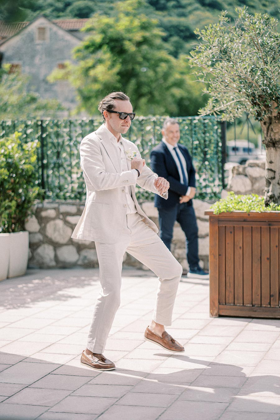 Man in a light beige suit and sunglasses dancing energetically outdoors on a sunny day, with a green garden and rustic stone wall in the background.