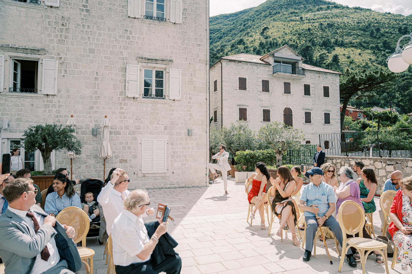 Outdoor wedding ceremony with guests seated on a stone patio, surrounded by historic stone buildings and a scenic mountain backdrop.