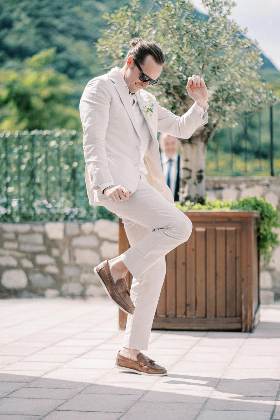 A man in a light beige suit joyfully dancing outdoors on a sunny day, with greenery and mountains in the background.