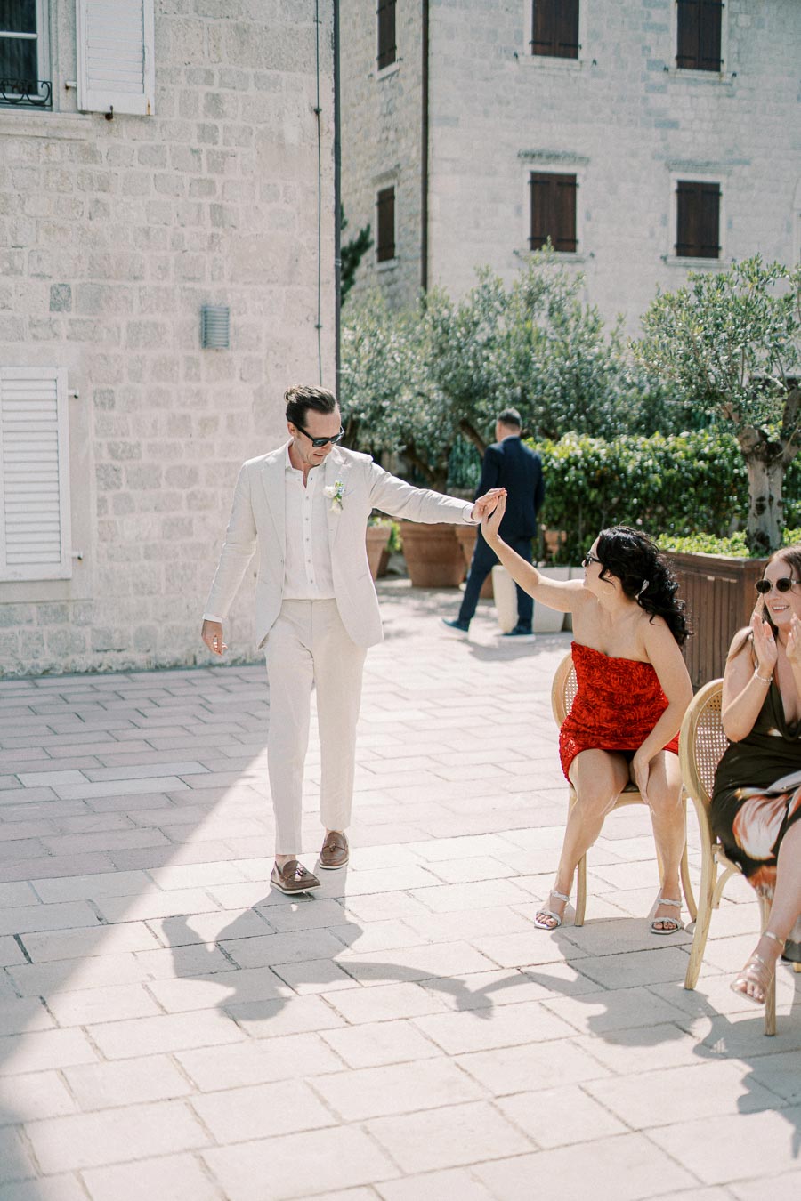 Elegant couple sharing a dance in a sunlit courtyard, man in light suit and woman in red dress, surrounded by stone buildings and greenery.