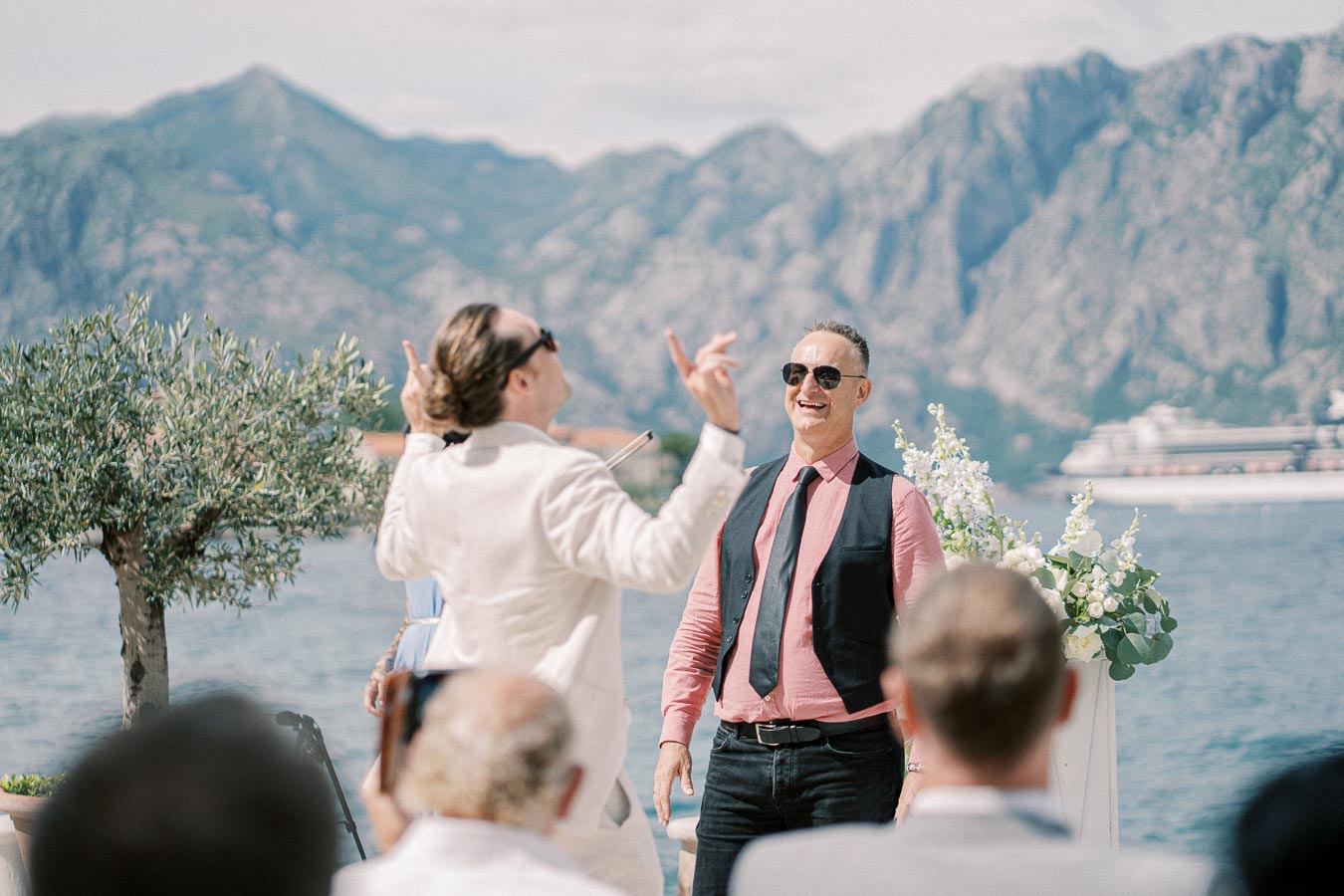 Outdoor event by the sea with two men smiling, dressed in casual formal attire, mountains in the background, and a cruise ship visible in the distance.