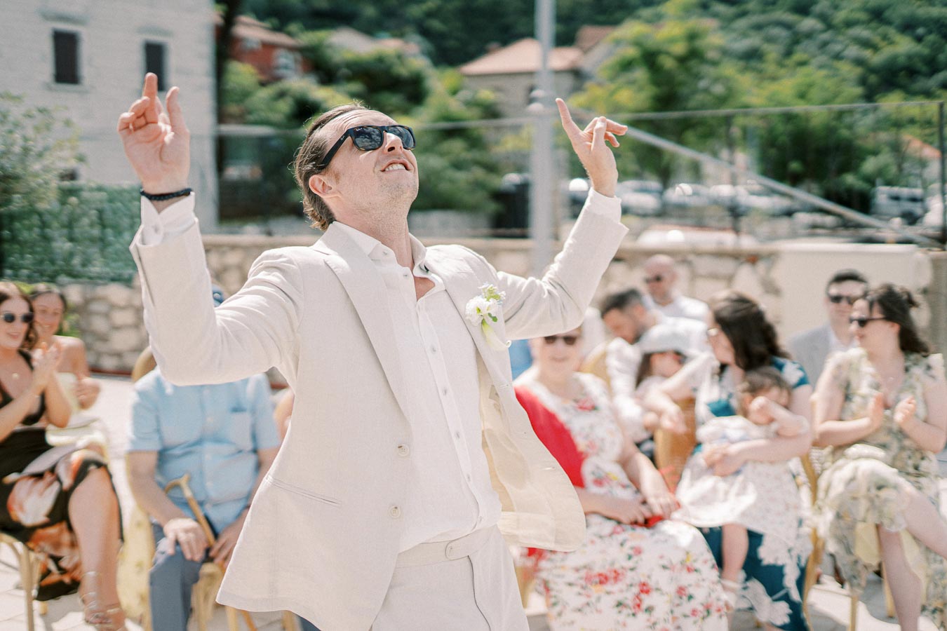A joyful man in a cream suit and sunglasses celebrating outdoors at a summer wedding, surrounded by smiling guests clapping in the background.