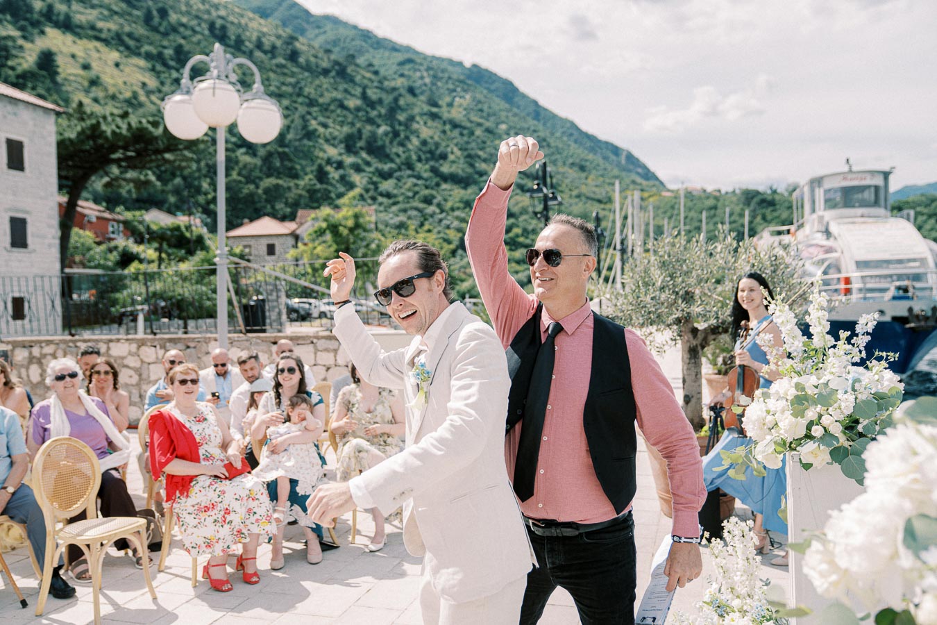 Two joyful men wearing sunglasses celebrate at an outdoor wedding ceremony, surrounded by seated guests and floral decorations, with a scenic mountain backdrop and a violinist performing in the background.