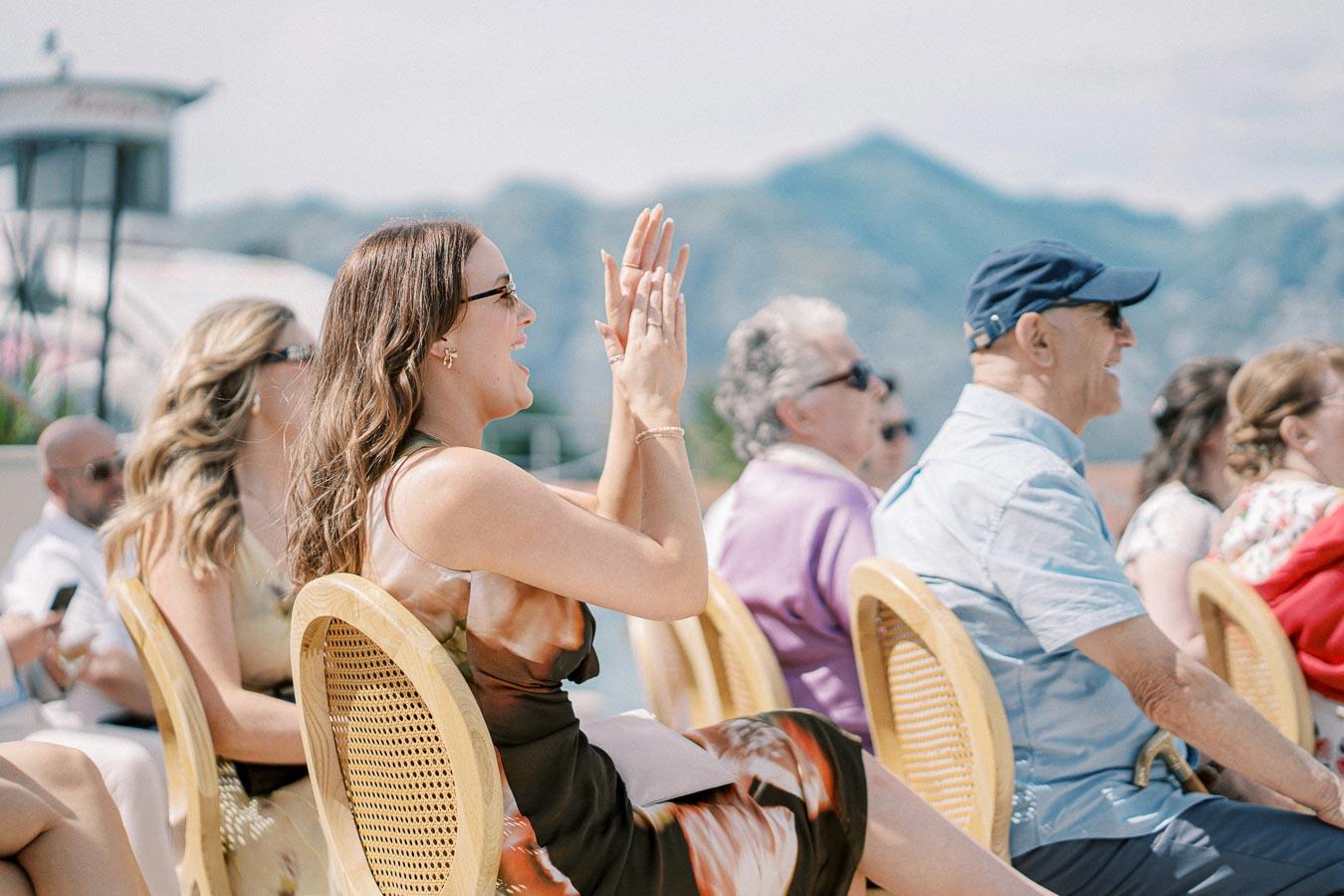 A group of people sitting outdoors and enjoying a sunny event, with mountains in the background. A woman in the foreground is clapping and smiling, wearing sunglasses and a summer dress.