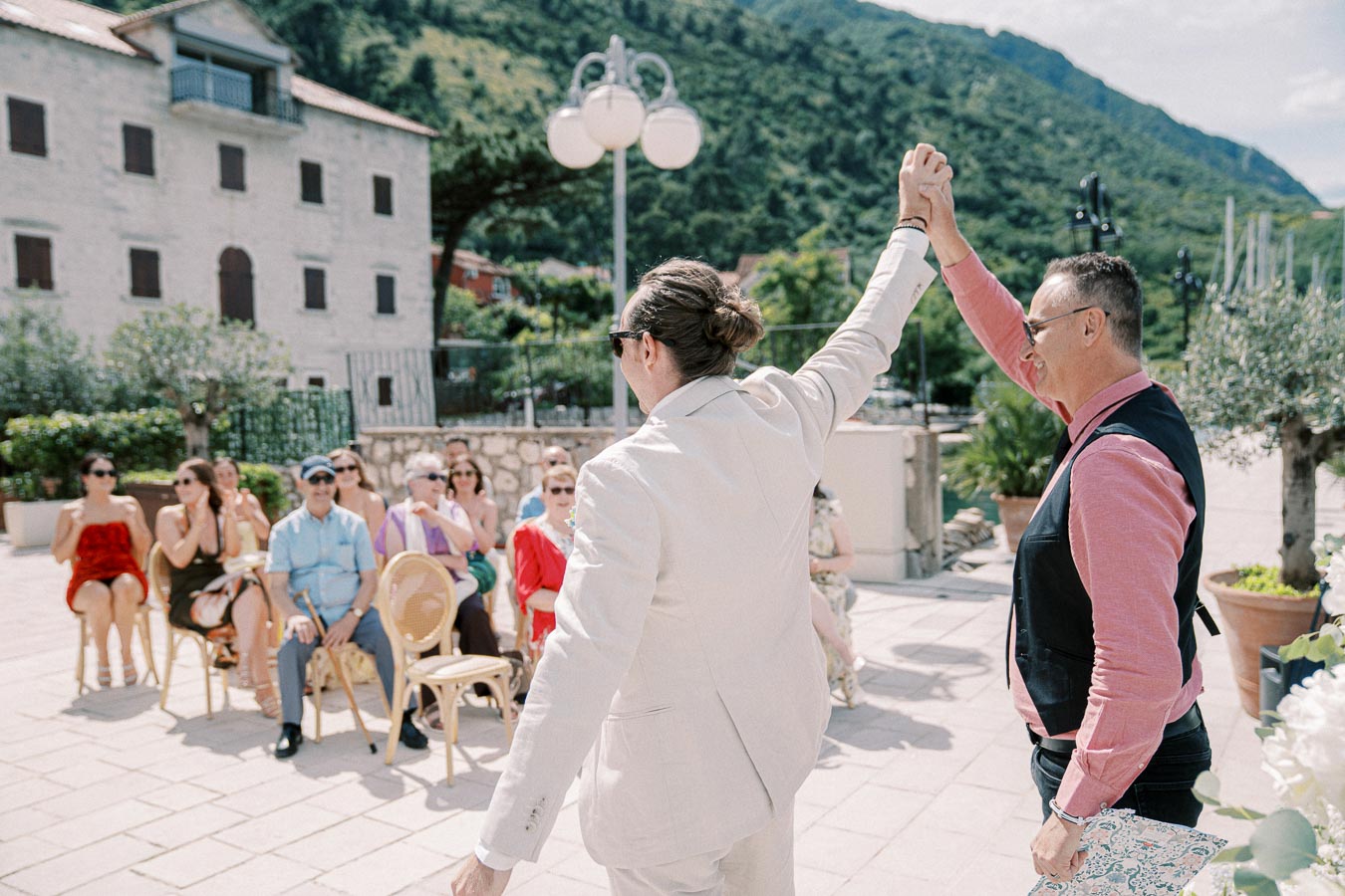 Outdoor wedding ceremony with two grooms celebrating, surrounded by seated guests and scenic mountain backdrop.