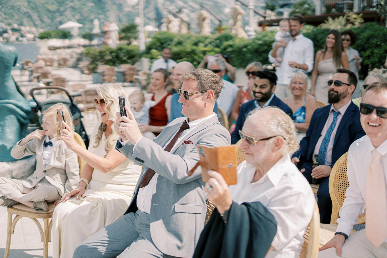 A group of well-dressed wedding guests sitting outdoors, smiling and taking photos with their phones, amidst a picturesque background of greenery and water, showcasing a joyful celebration.