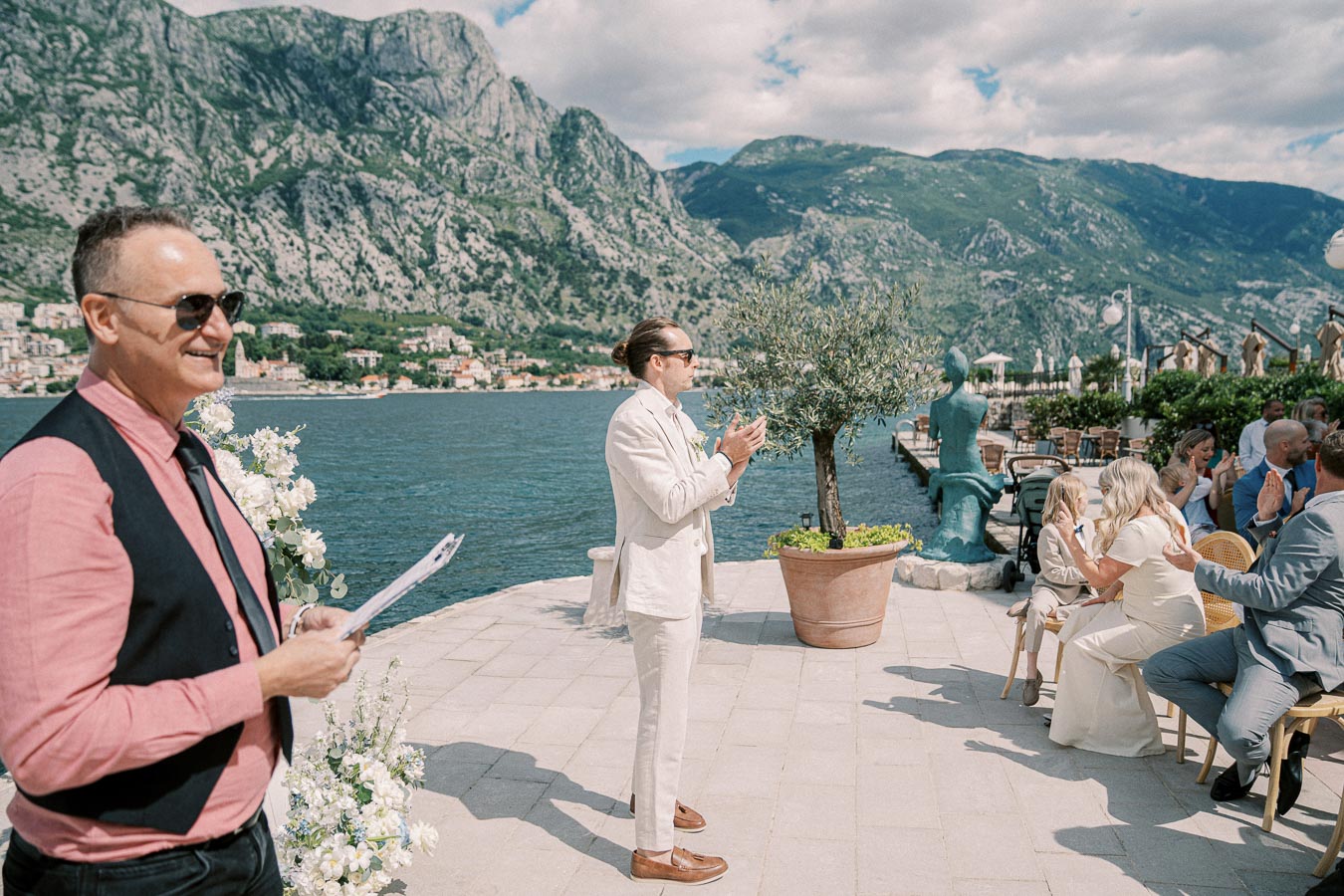 A wedding ceremony by the sea with a scenic mountainous backdrop. A man in a light suit stands clapping, while guests in elegant attire are seated on a sunny patio adorned with flowers. The atmosphere is joyful and serene, ideal for a picturesque destination wedding.