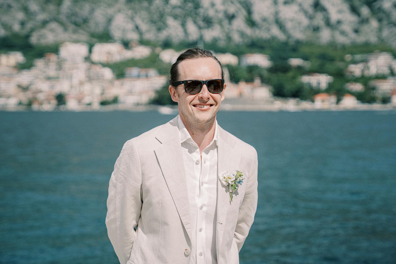 Man in a white suit with sunglasses smiling by a lake with a city and mountains in the background.