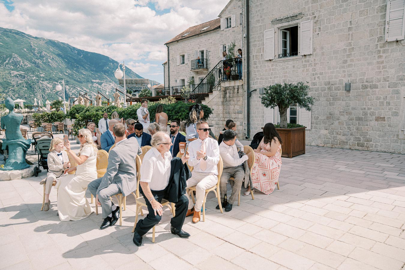 A group of people attending an outdoor event on a sunny day at a rustic stone venue with scenic mountain views. They are seated on wooden chairs, engaged in conversation, with a backdrop of lush greenery and clear blue skies.