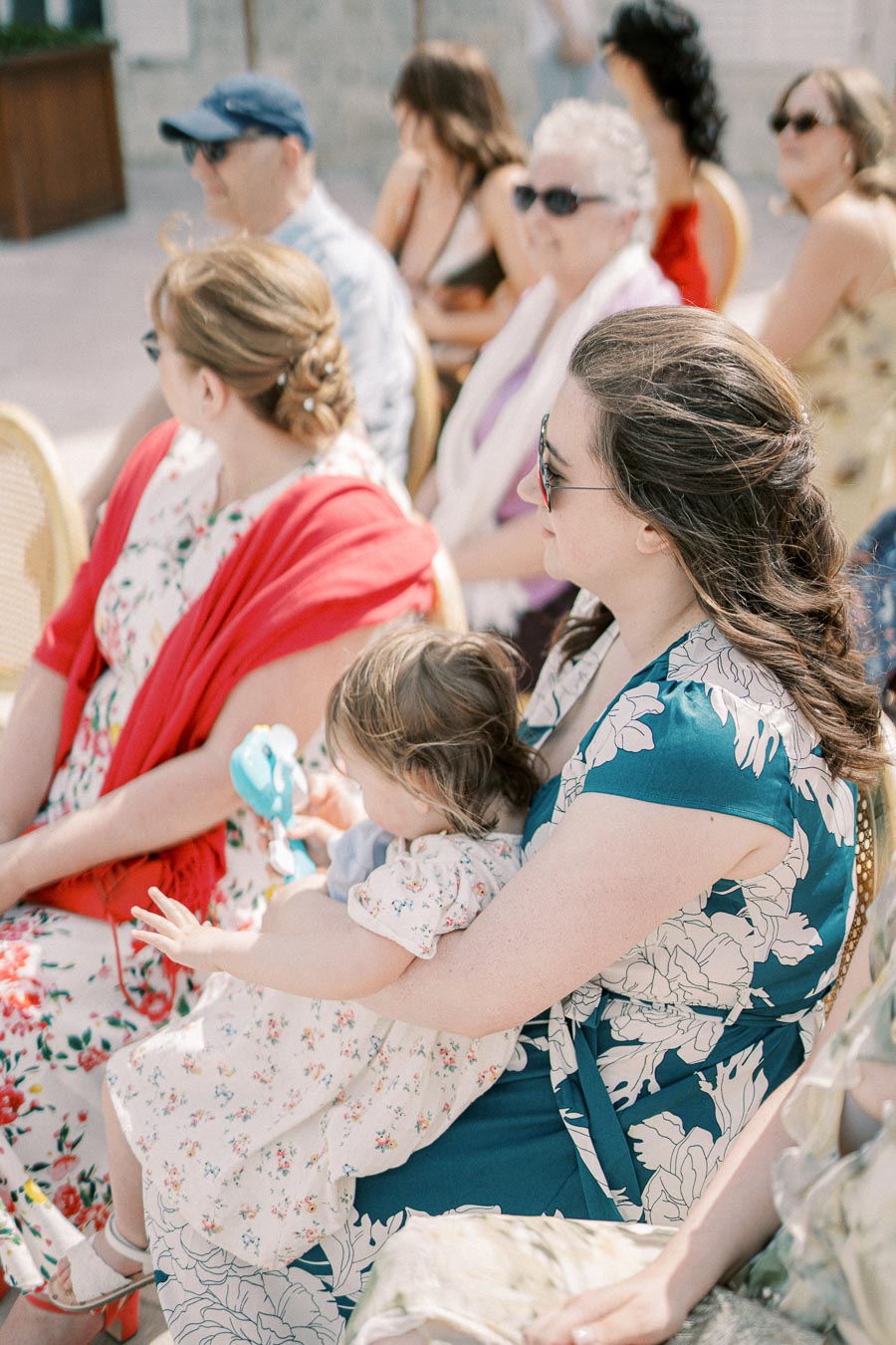 A group of people sits at an outdoor event, with a woman holding a child on her lap. The attendees are dressed in floral patterns and light clothing, suitable for a sunny day.