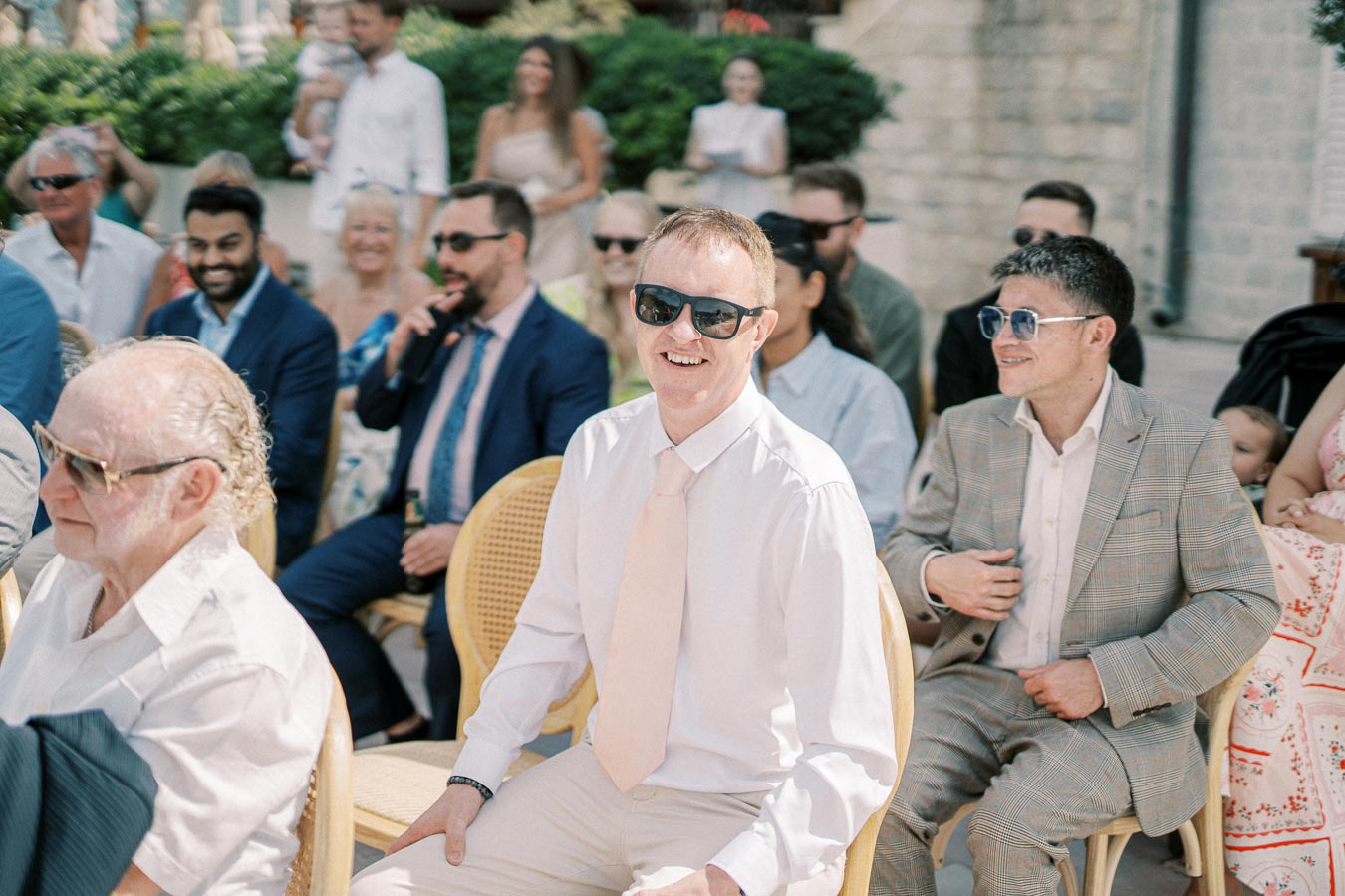 A group of people seated outdoors at a wedding ceremony, dressed in formal attire with sunglasses, enjoying the sunny weather.
