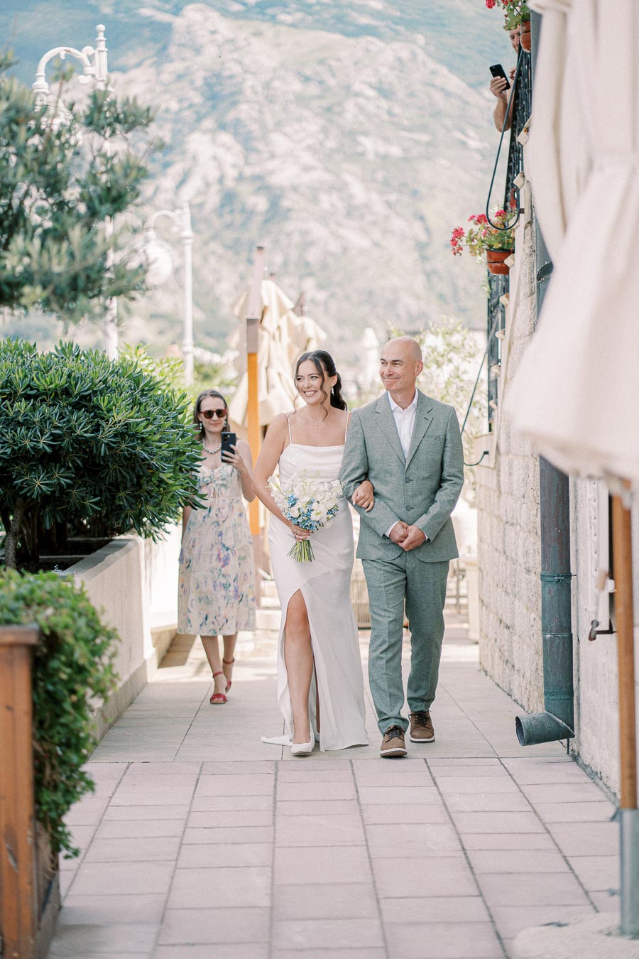 A bride in a white wedding dress walking with a man in a light suit down a picturesque path, with mountains in the background and guests taking photos.